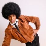 Mężczyzna z dużą fryzurą afro w zamszowej kurtce i krawacie.
